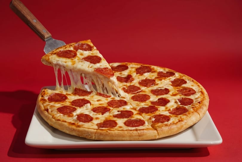 A pepperoni pizza on a white plate with a slice being lifted, showing melted cheese stretching.