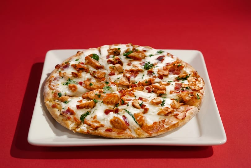 A round flatbread pizza with melted cheese, chicken, bacon, and herbs on a white plate.