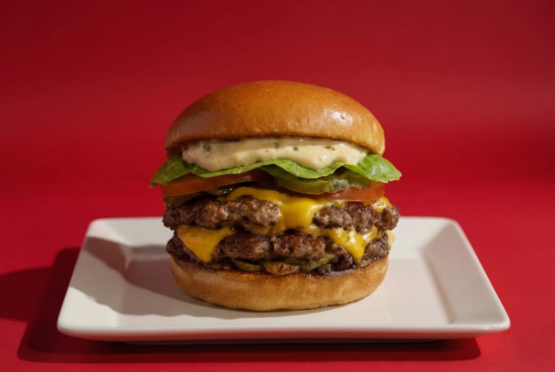 Double cheeseburger with lettuce, tomato, pickles, and sauce on a white plate against a red background.