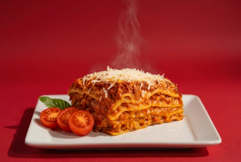 Steaming hot lasagna with grated cheese, basil, and sliced tomatoes on a white plate.
