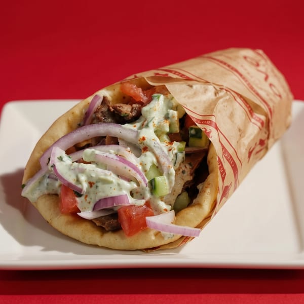 A gyro wrapped in paper, topped with tzatziki, red onions, and tomatoes, on a white plate.