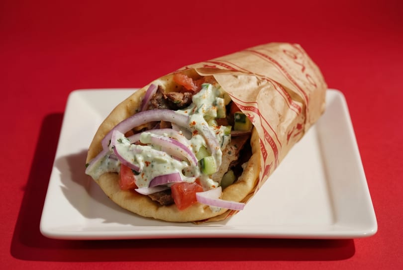 A gyro wrapped in paper, topped with tzatziki, red onions, and tomatoes, on a white plate.